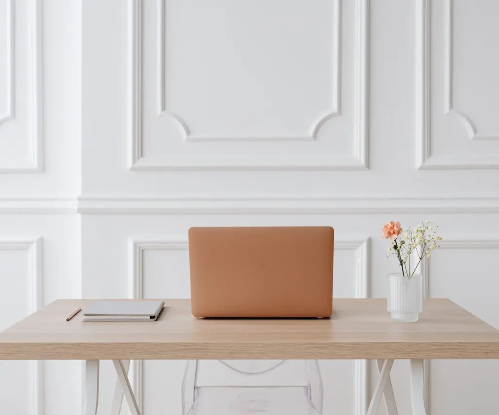 Scrivania in legno chiaro con quaderno e portatile, davanti a una parete a boiserie bianca